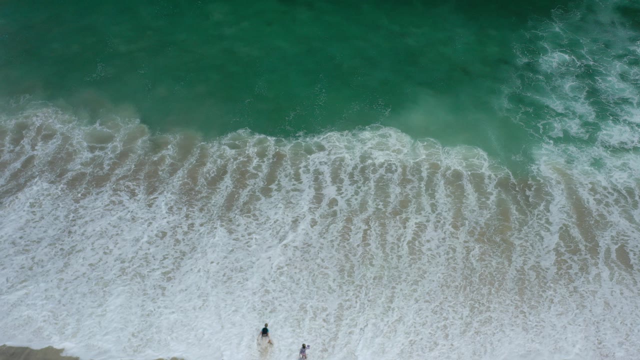 Overhead green surf breaking onto sand with tiny people near the lower edge.