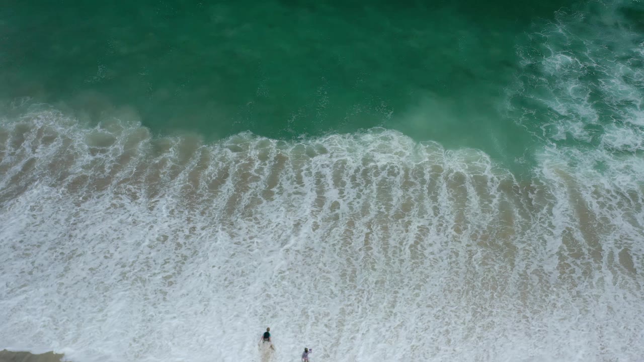 Green surf and white foam cover the Los Cabos shoreline from above, with tiny figures still near the lower edge and no horizon in view.