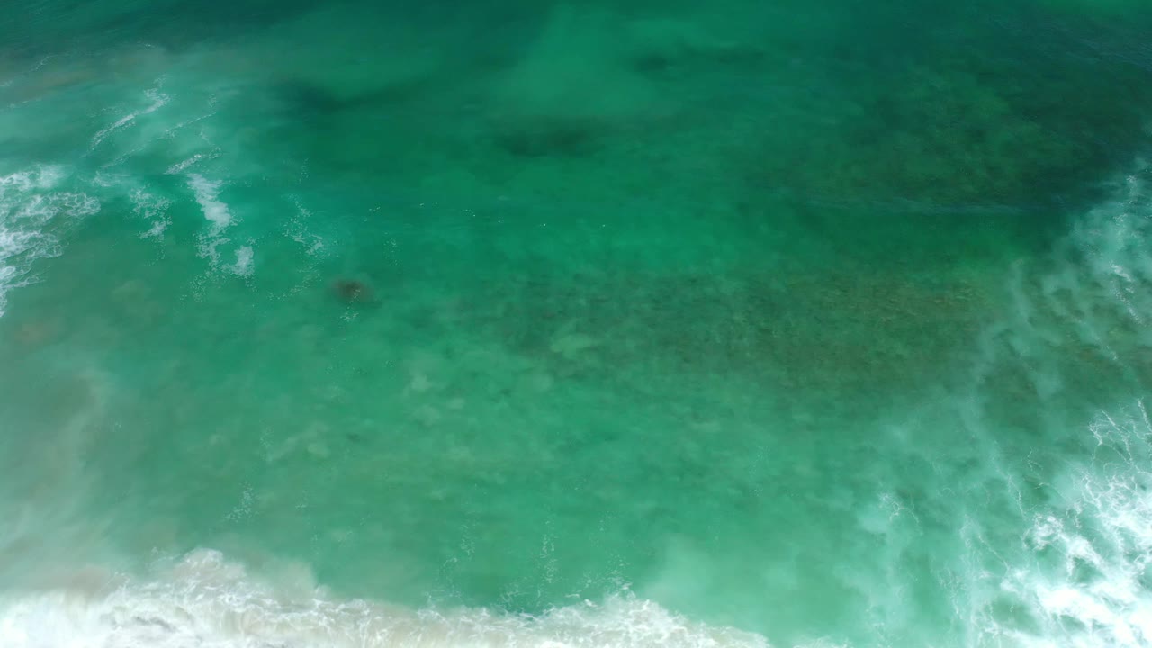 Overhead Los Cabos water view with a broader white foam streak crossing the aqua surface and darker underwater areas visible, no visible people.