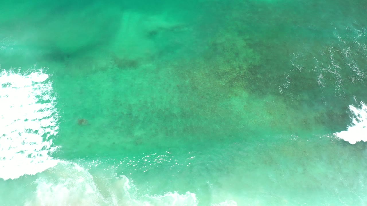 Overhead Los Cabos ocean frame with a bright white wave breaking near the center and darker underwater patches visible around it, no visible people.