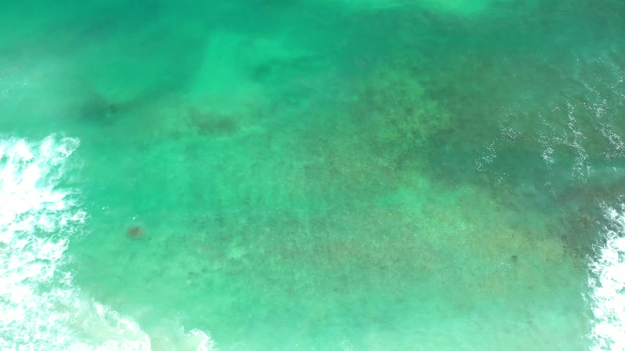 Aerial frame of clear Los Cabos ocean water with pale aqua shallows, darker underwater patches, and a narrow white wave streak near the lower center, with no visible people.