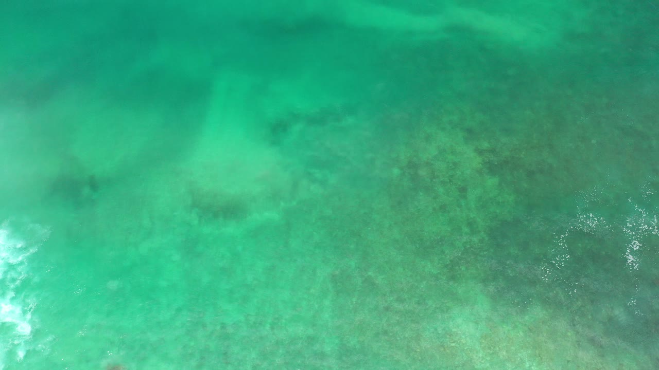 Overhead view of green ocean water in Los Cabos, Mexico, with darker submerged reef texture and small white foam near the lower edge, no visible people.