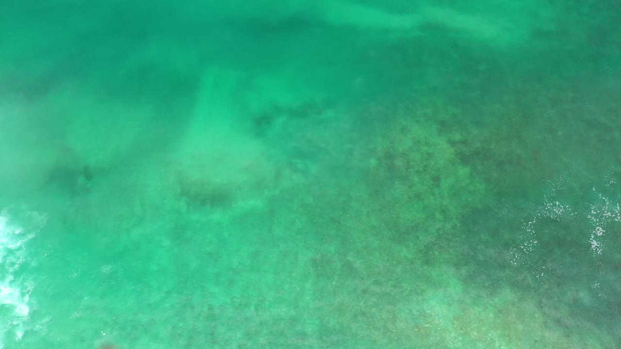 Overhead Los Cabos ocean frame with translucent green water, darker reef below the surface, and a bright white foam streak toward the center-right, no visible people.
