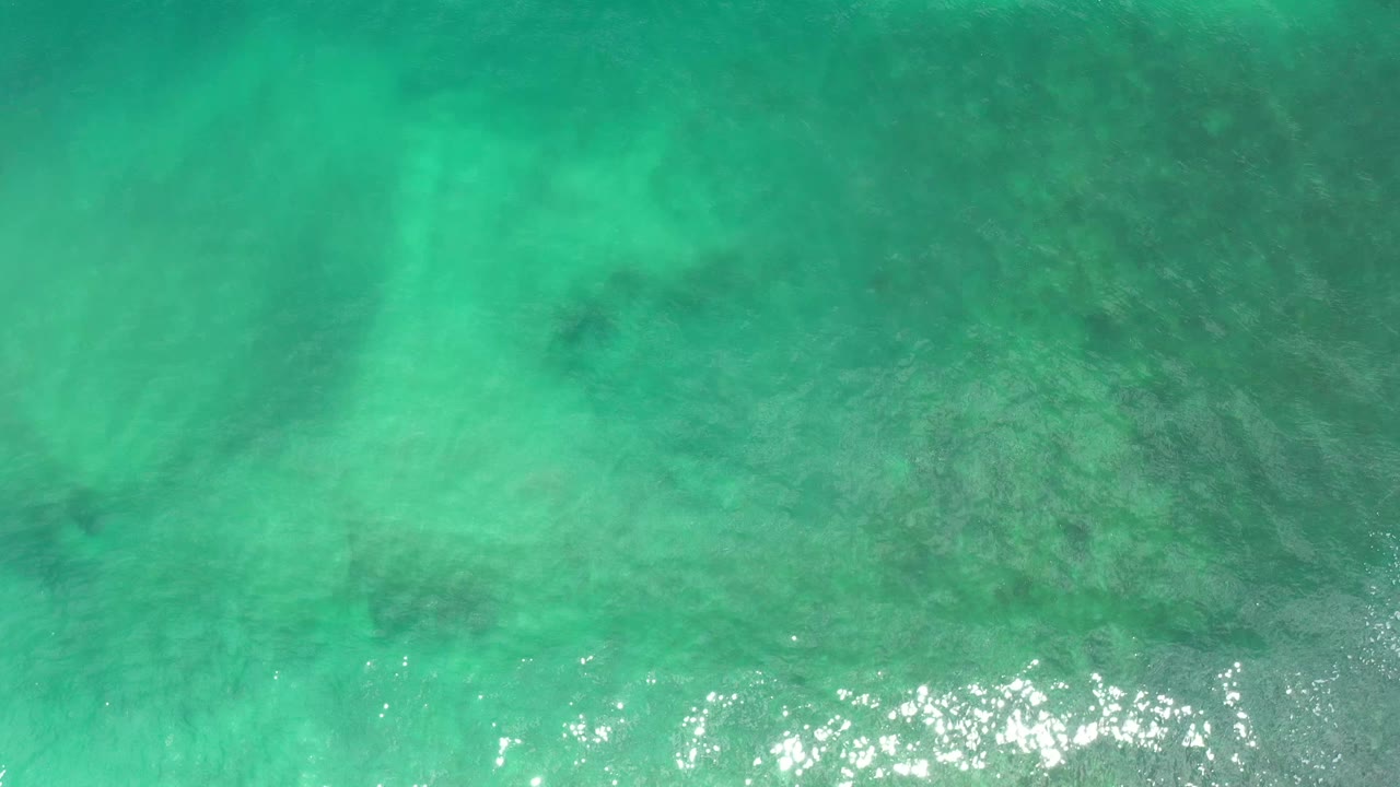 Clear turquoise ocean water in Los Cabos, Mexico, seen from above with scattered dark patches under the surface and no visible people.