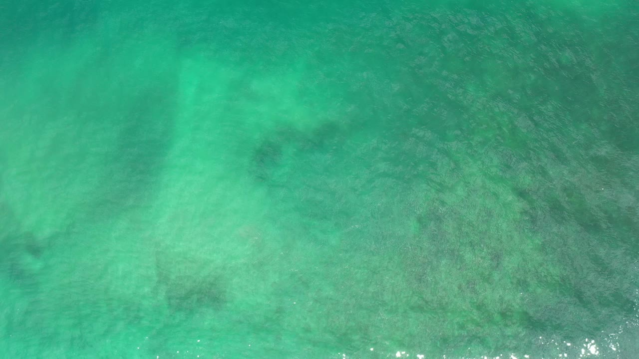 Aerial ocean water with pale green areas, darker submerged texture, and bright reflections along the lower edge.