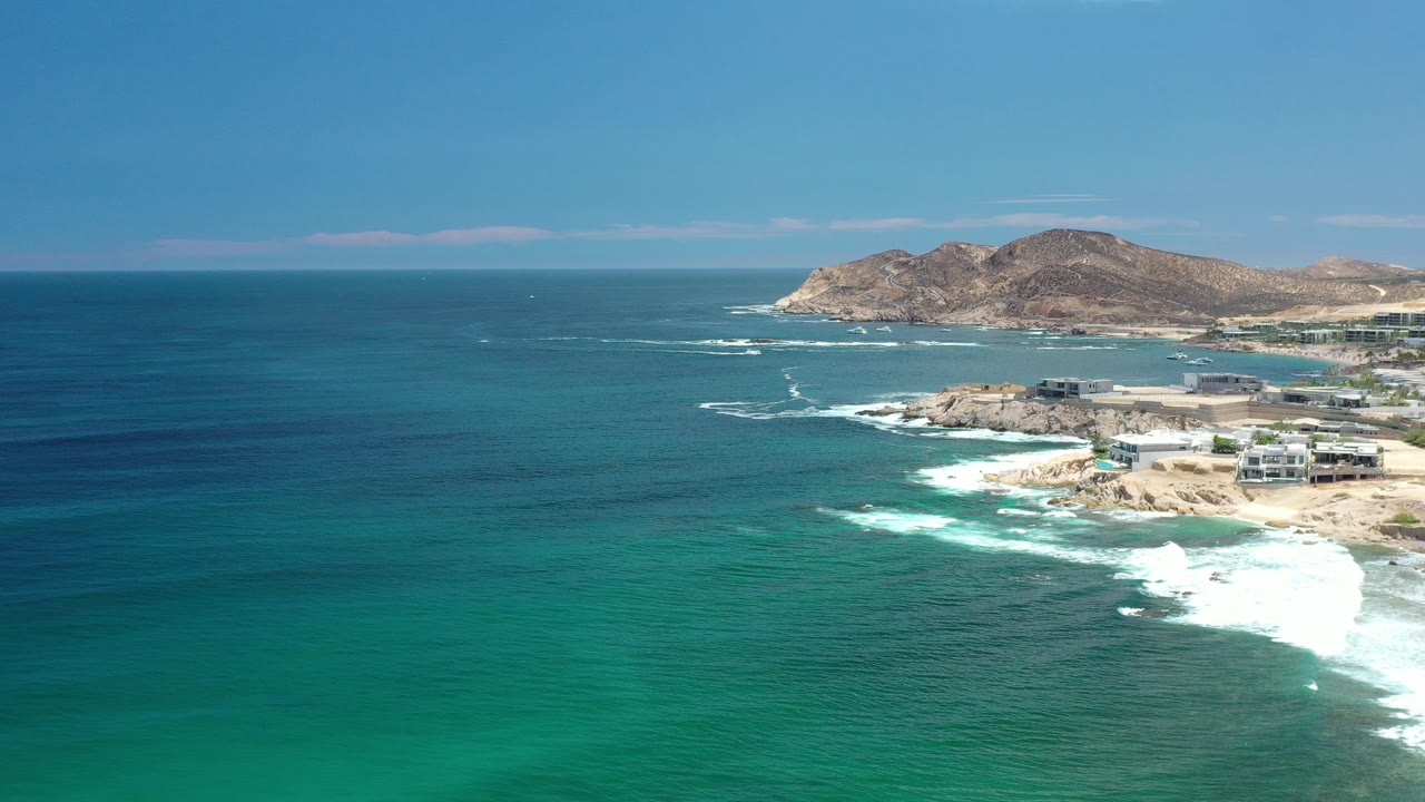 Aerial view of blue-green ocean, surf, coastline, buildings, and hills.