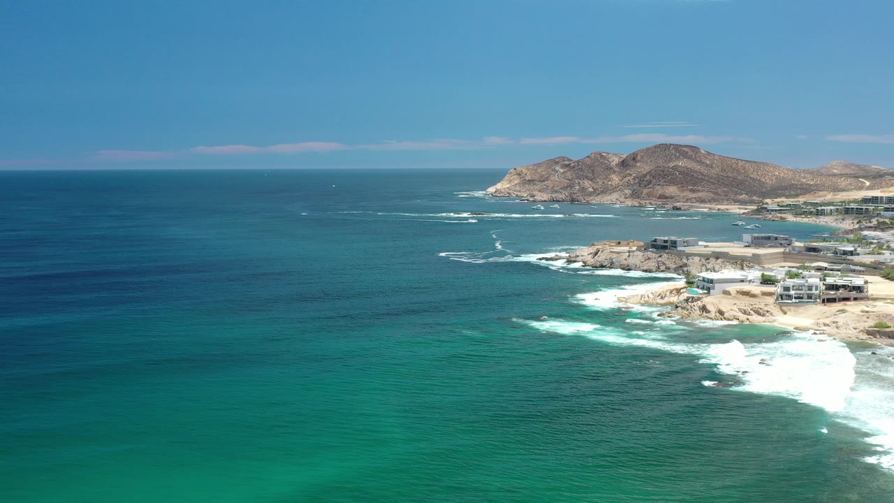 Aerial view over the Los Cabos coastline where the ocean dominates the foreground, surf wraps the shore, buildings sit near the beach, hills line the background, and no visible people appear.