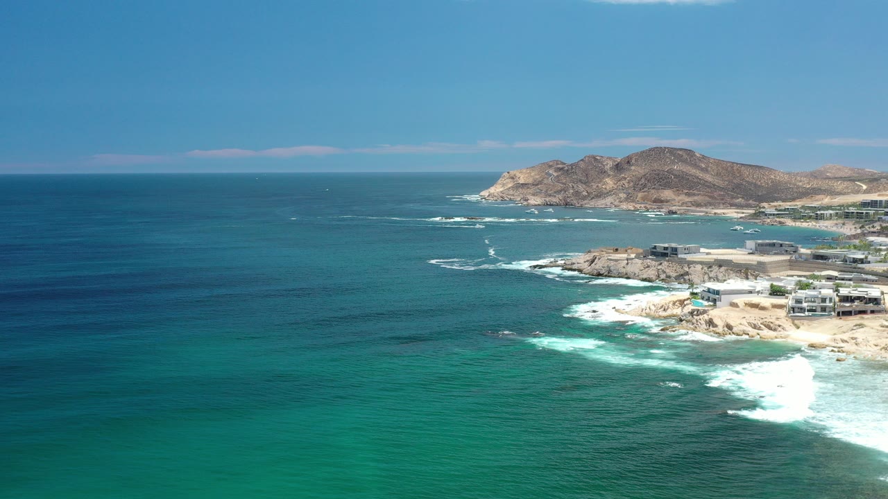 Aerial view over Los Cabos, Mexico with turquoise water in the foreground, white surf breaking along the rocky sandy shoreline, distant hills, and no visible people.