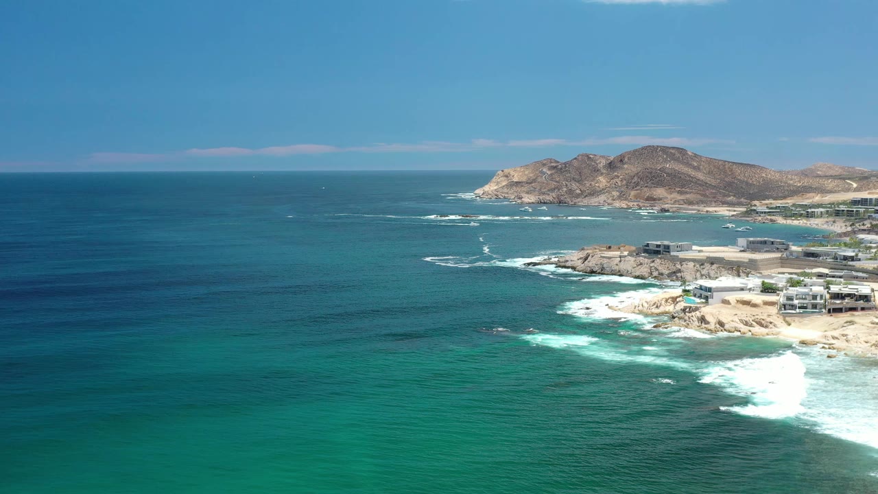 Wide Los Cabos coastal aerial showing deep blue water shifting to turquoise near shore, white waves along the beach, low mountains beyond, and no visible people.
