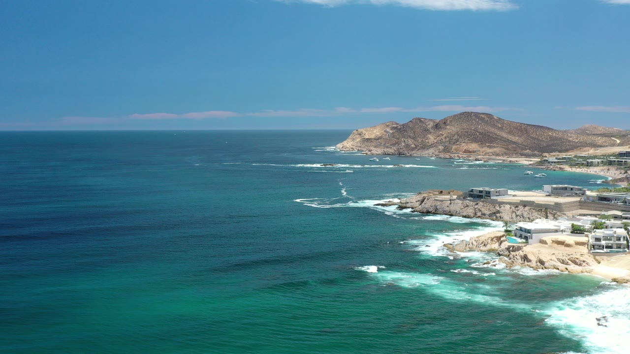 Opening aerial view of the Los Cabos coastline with deep blue ocean, turquoise shallows, waves breaking on rocks, dry hills beyond, and no visible people.