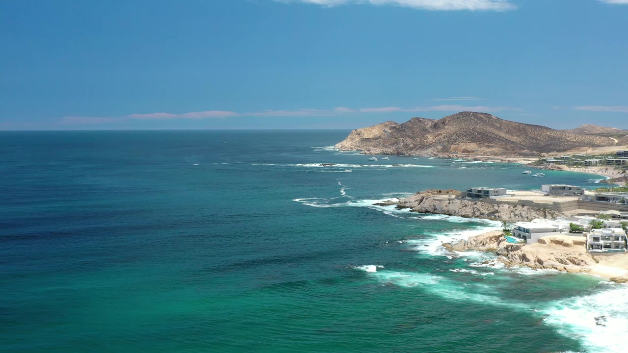 Aerial Los Cabos shoreline view with a dark blue water band offshore, white surf curling around tan rocks, buildings at the right edge, and no visible people.