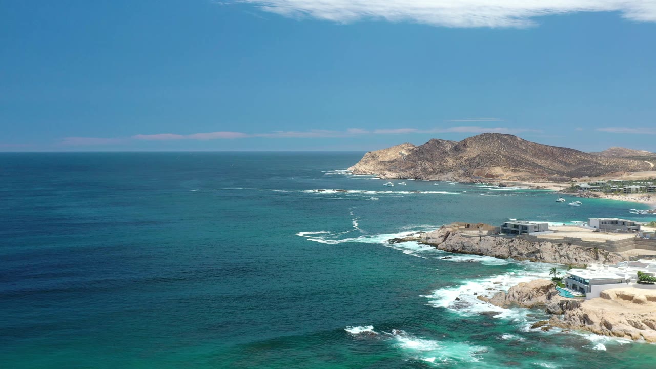 Aerial frame over Los Cabos, Mexico, with a broad stretch of blue water at left, white wave lines near the rocks, low brown islands beyond, and no visible people.