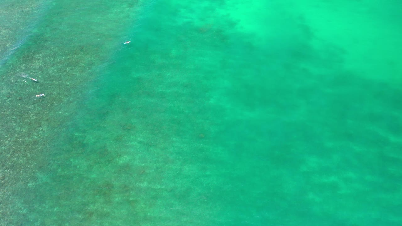 Overhead view of turquoise-green ocean in Los Cabos, Mexico with darker reef texture at left, tiny white watercraft near the upper left, and no visible people.