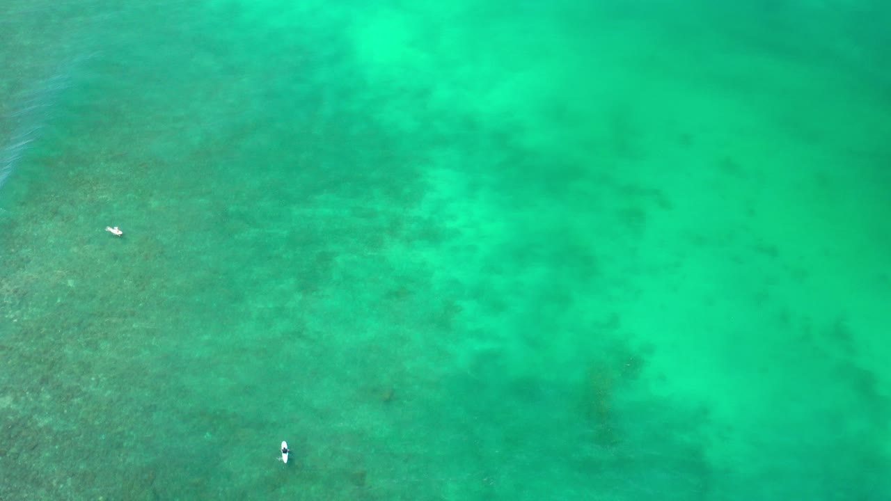 Overhead view of turquoise water in Los Cabos, Mexico, with darker submerged patches, faint ripples, tiny pale floating shapes, and no visible people.