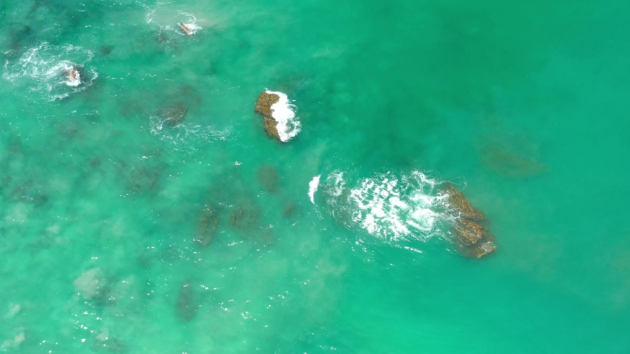 Overhead view of turquoise water in Los Cabos, Mexico, with scattered submerged rocks, one exposed rock, small white foam patches, and no visible people.