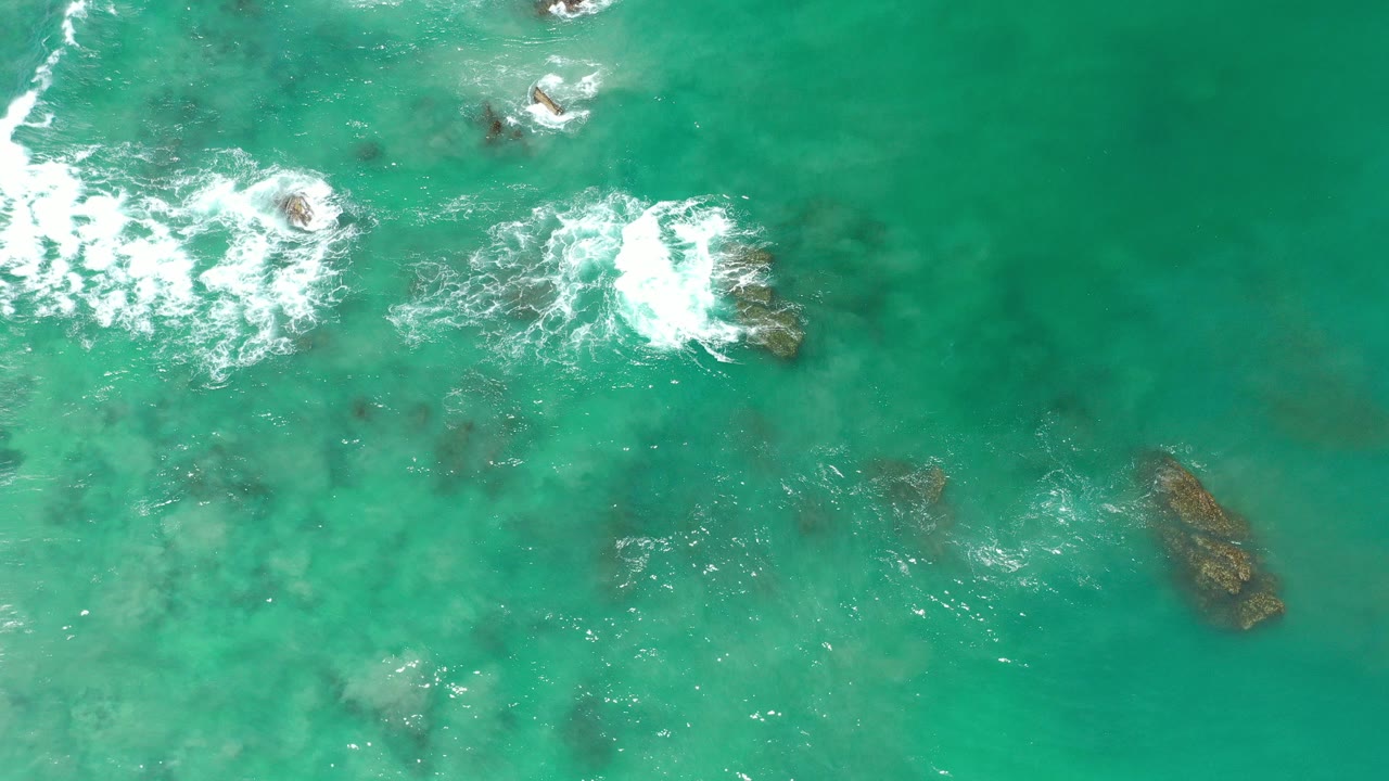 Overhead view in Los Cabos, Mexico, of turquoise shallow water with white foam spreading over dark reef rocks, no visible people.