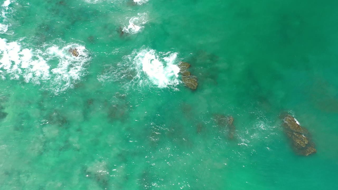 White wave bursts around rocks in teal shallow ocean water.