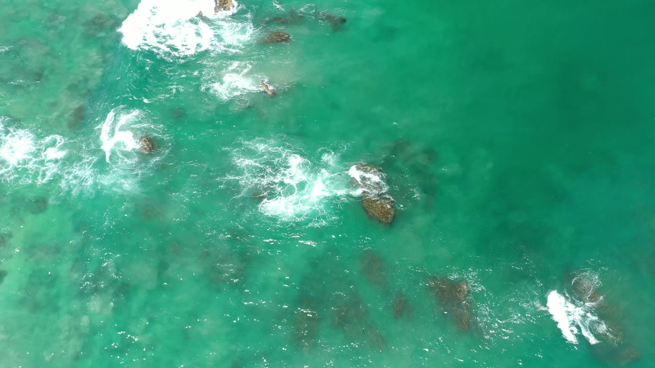 Aerial Los Cabos ocean frame with teal water, bright white foam curling around several dark rocks, and no visible people or shoreline.