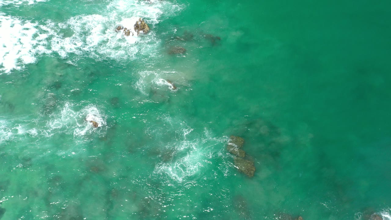 Overhead view of turquoise ocean water in Los Cabos with white surf breaking near scattered dark rocks, no visible people.