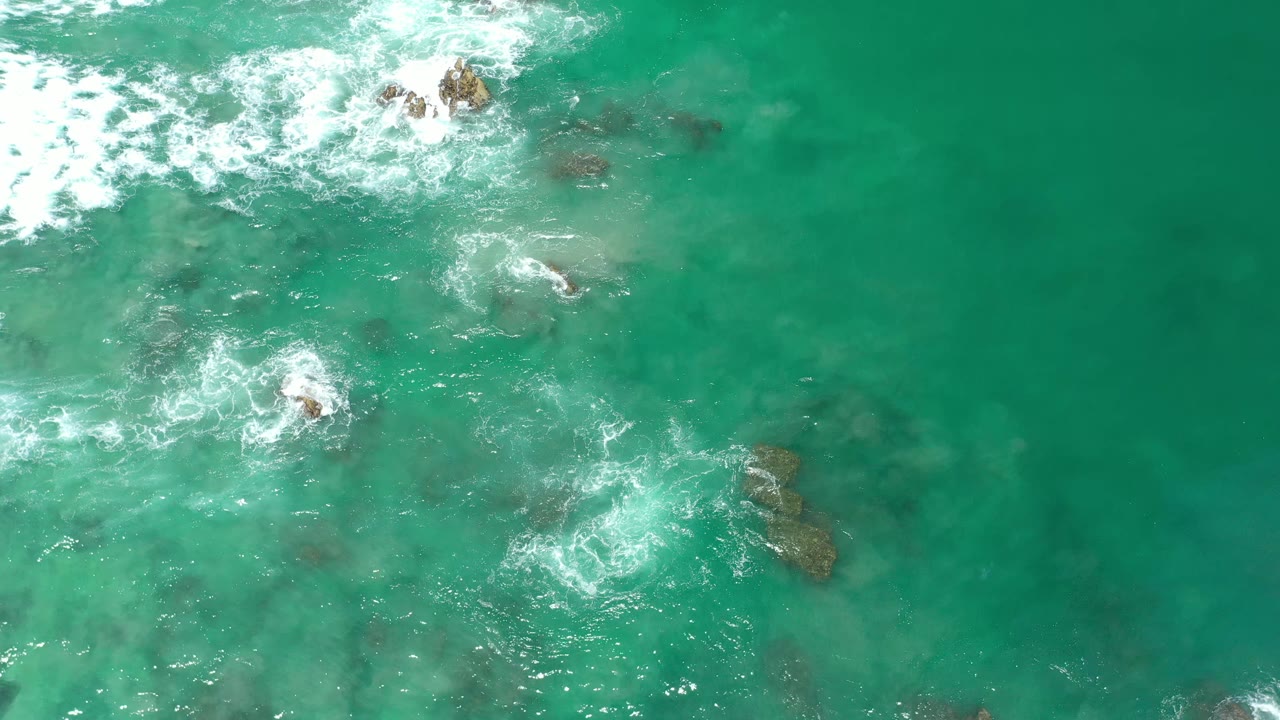 Overhead Los Cabos water view with foamy surf widening across clear green-blue water and dark rocks below, no visible people.