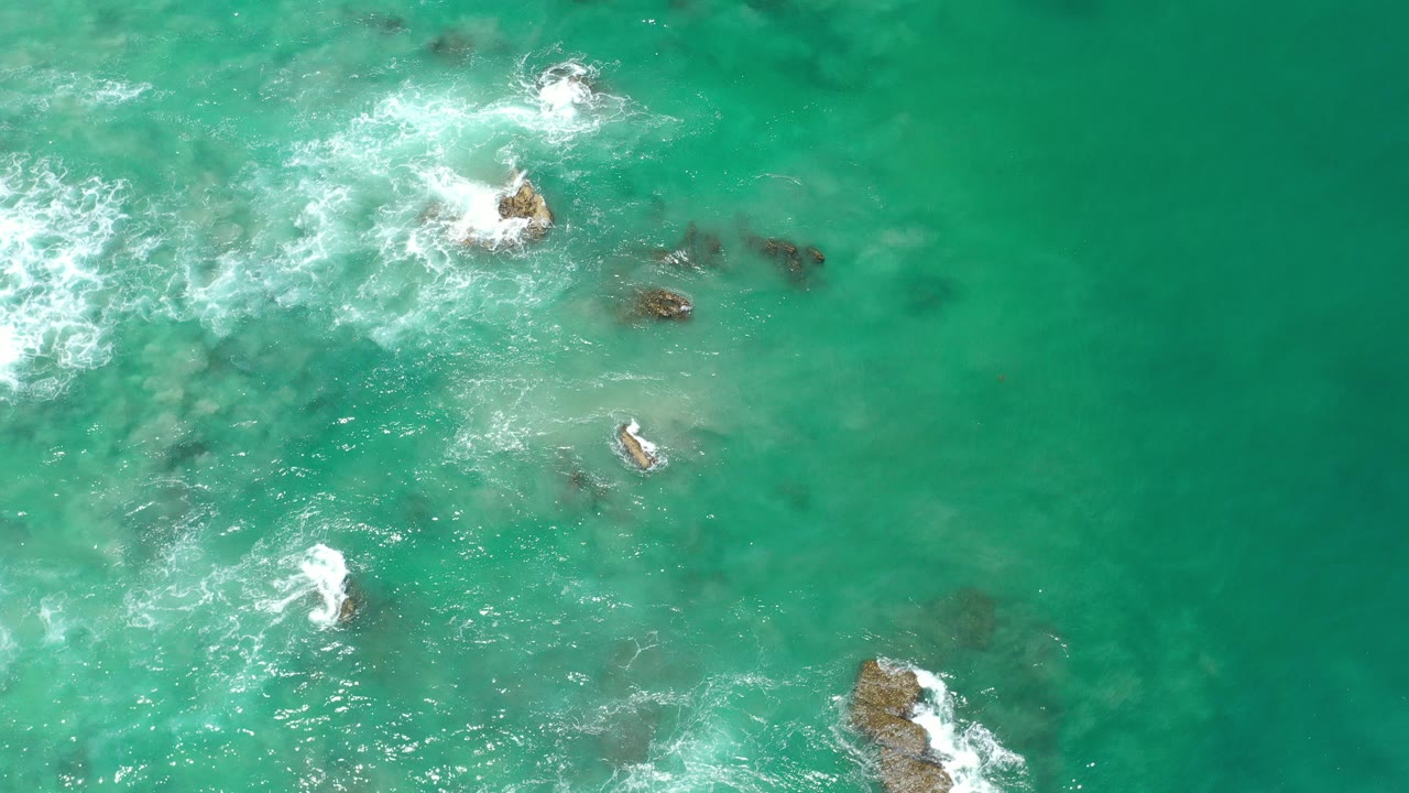 Top-down view of turquoise water in Los Cabos, Mexico, with white foam around several rocks and no visible people.