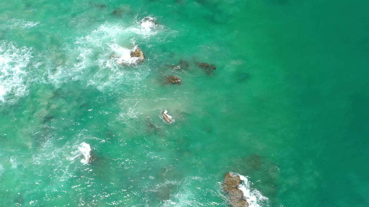 Aerial frame over Los Cabos water where white surf curls around central rocks, with darker underwater shapes and no visible people.