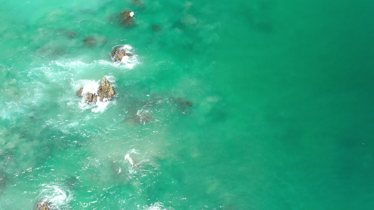 Top-down aerial view in Los Cabos, Mexico, showing turquoise seawater, scattered rocks near the left side, white surf around exposed points, and no visible people.