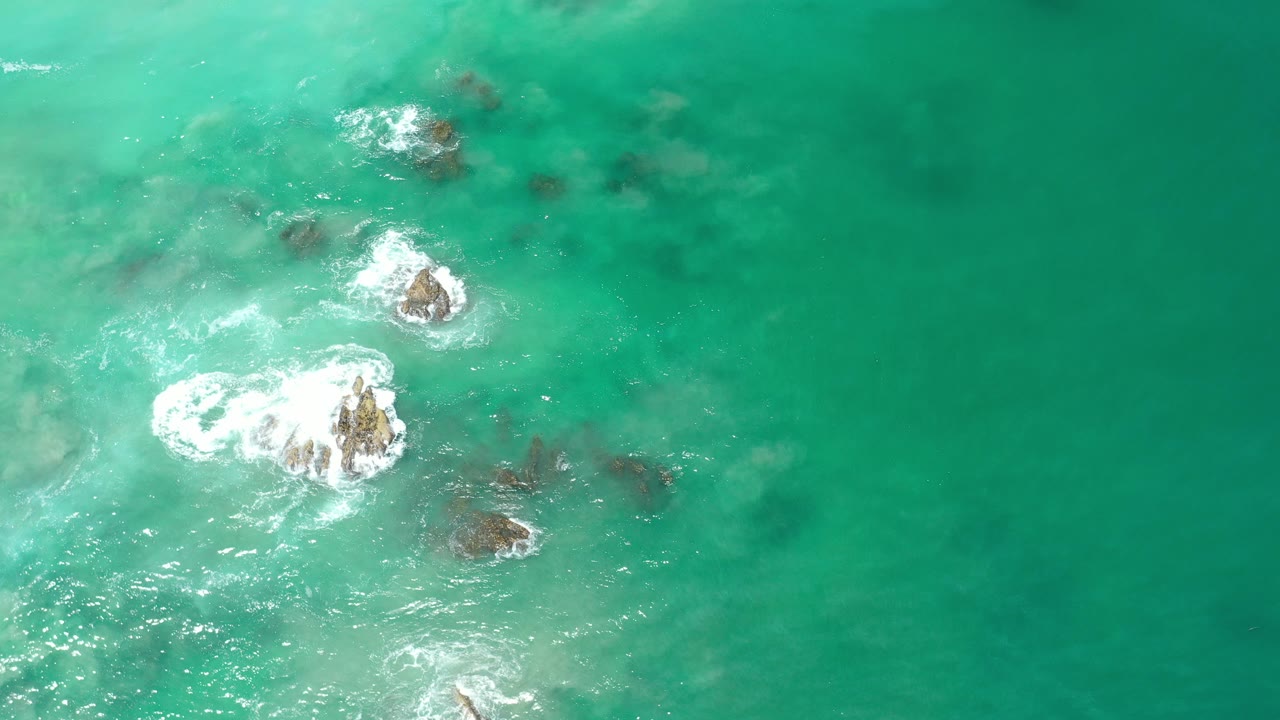 Overhead view of turquoise water in Los Cabos, Mexico, with several rocks breaking the surface, white foam around the lower-left cluster, and no visible people.