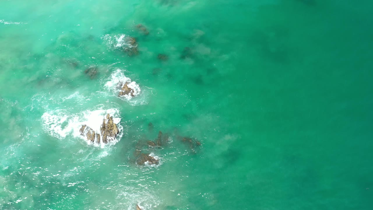 Rock clusters in green-blue water with waves breaking around them.