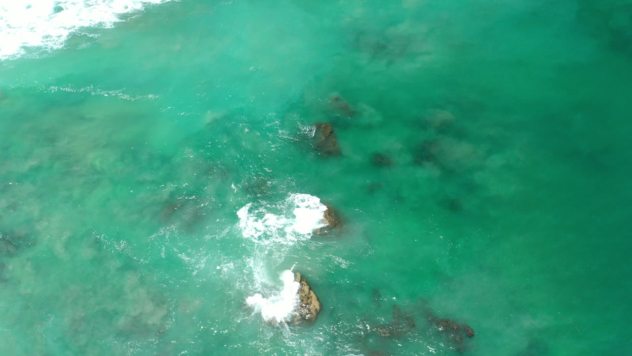 Overhead view of green ocean water in Los Cabos, Mexico, with white foam breaking around several dark rocks and no visible people.