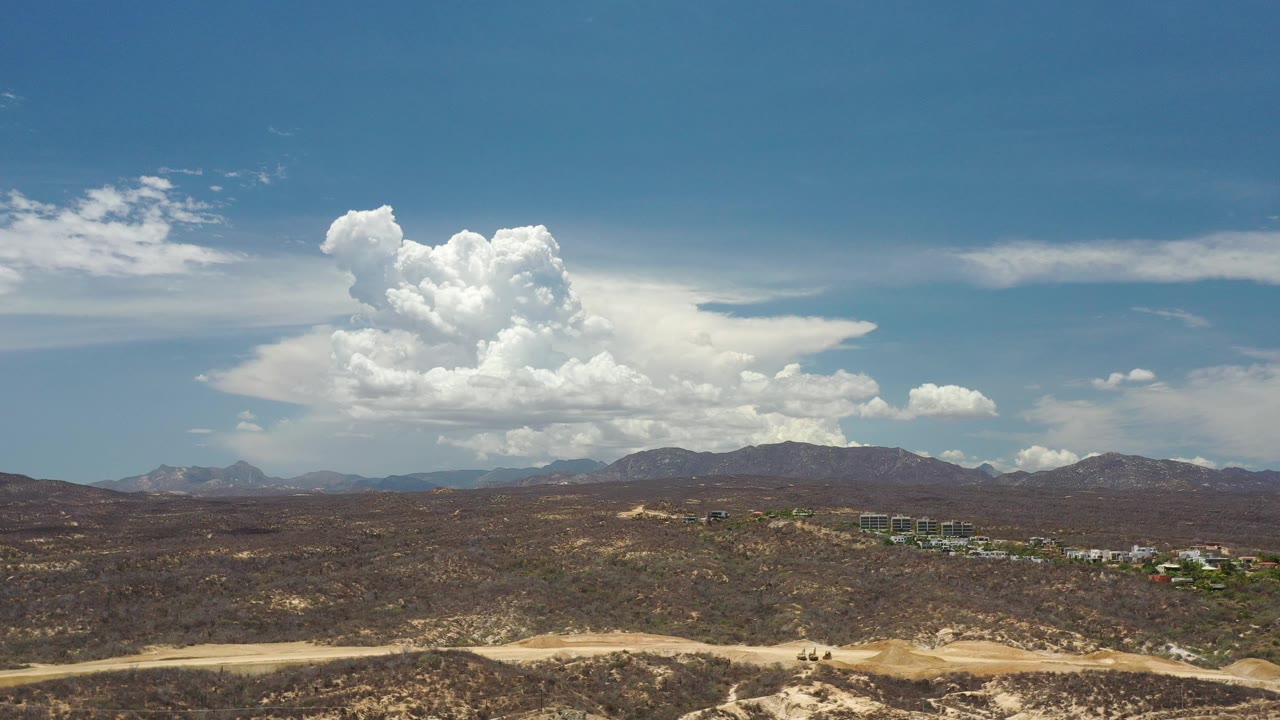 Opening frame of Los Cabos, Mexico, showing a highway at the bottom, arid scrubland, distant mountains, and a large cloud bank under blue sky with no visible people.
