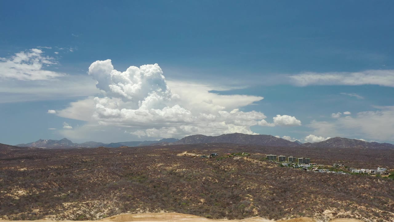 Los Cabos desert ridges stretch across the frame under a bright cumulus cloud mass, with small structures near the horizon and no visible people.