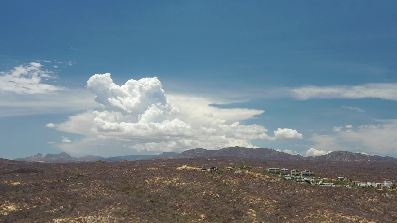 Wide view of Los Cabos desert terrain with distant mountains, a small building cluster, large white clouds, and no visible people.
