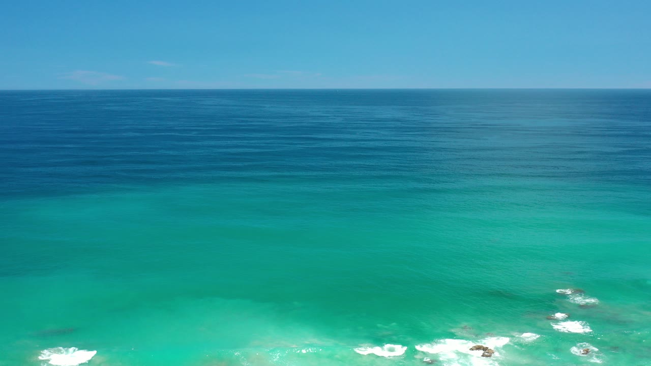 Aerial view over Los Cabos, Mexico water with turquoise shallows, deeper blue sea at the horizon, small white surf near the lower edge, and no visible people.