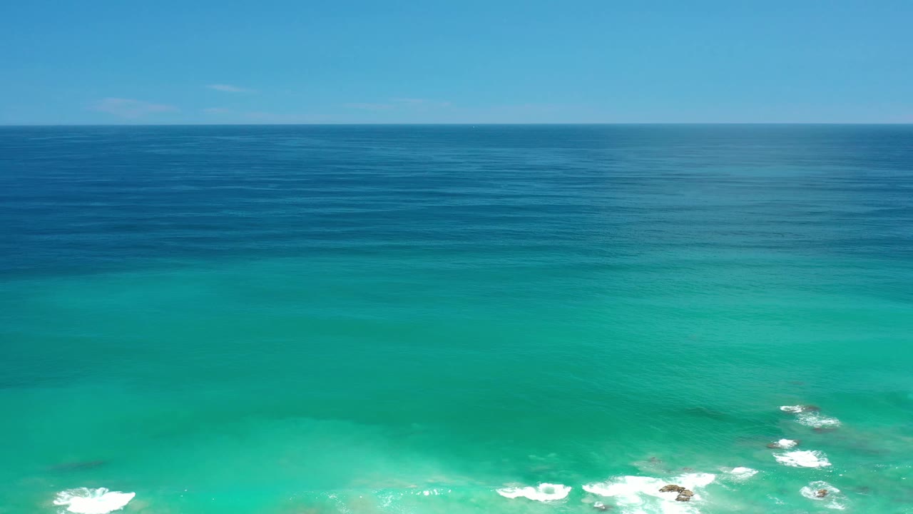 Los Cabos, Mexico aerial frame showing broad turquoise water in the foreground, darker blue ocean beyond, a level horizon, scattered reef wash, and no visible people.