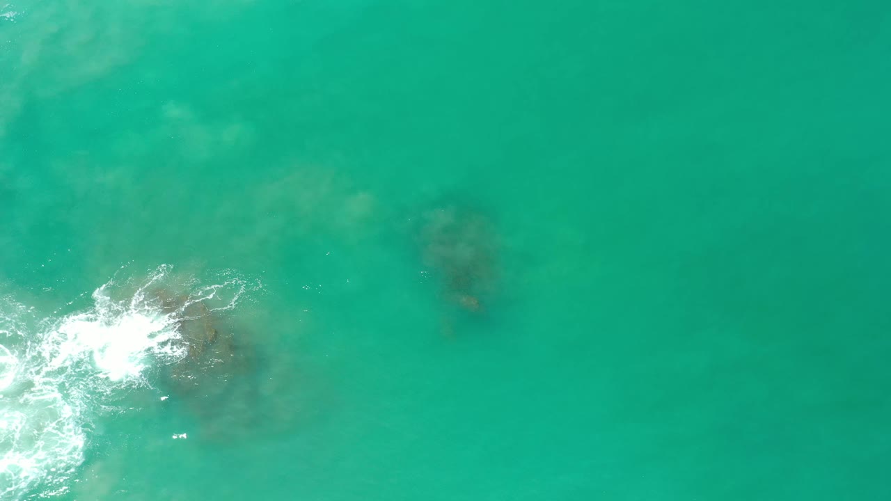 Aerial frame from Los Cabos, Mexico showing white water spreading across a central rocky patch surrounded by clear turquoise ocean, no visible people.