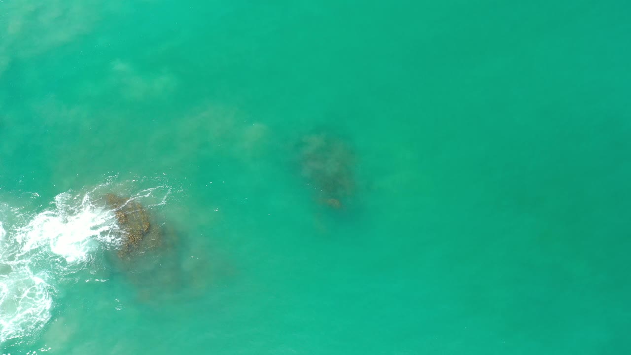 Foam curls around rocks near the left side of an overhead ocean view.