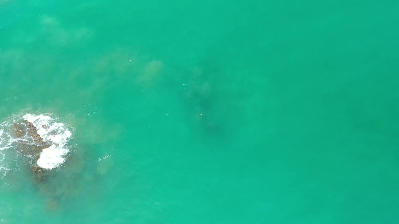 Aerial Los Cabos frame with foam spreading across the top of a submerged rock in turquoise green water, with no visible people.