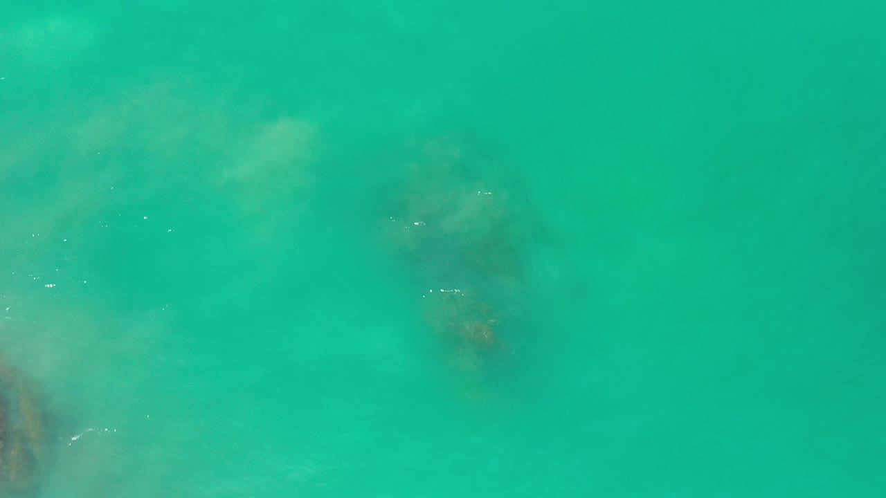 Overhead view of clear green water in Los Cabos, Mexico with hazy submerged darker patches near the left edge and no visible people.