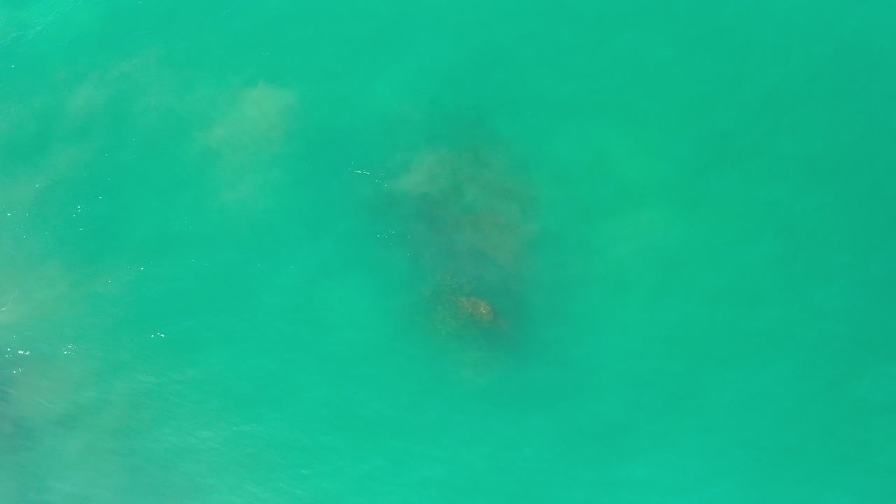 Overhead view of sea-green water in Los Cabos with a dark submerged rock shape under cloudy wave wash and no visible people.