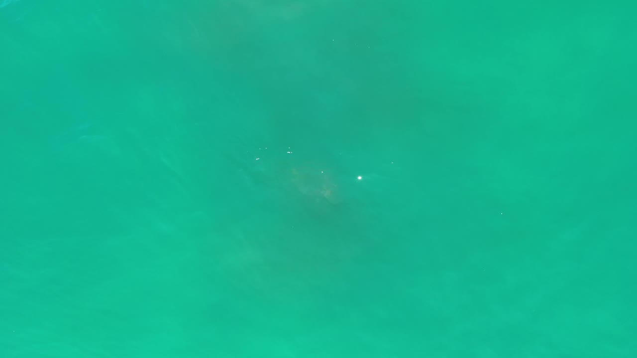 Aerial frame of turquoise water in Los Cabos, Mexico, showing small sunlit specks, soft darker areas below the surface, and no visible people.