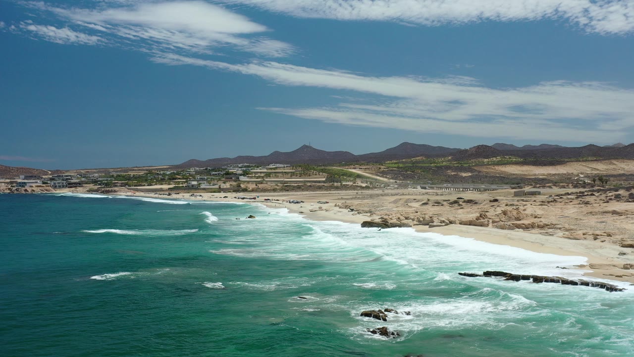 Opening aerial frame over a Los Cabos beach, with turquoise water in the foreground, white surf on sand, desert flats behind the shore, distant mountains, and no visible people.