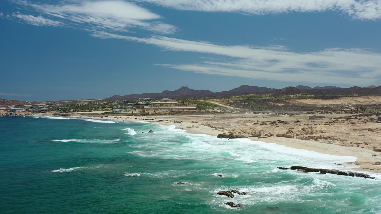 Wide coastal aerial with teal water, whitewater, beach, and desert hills