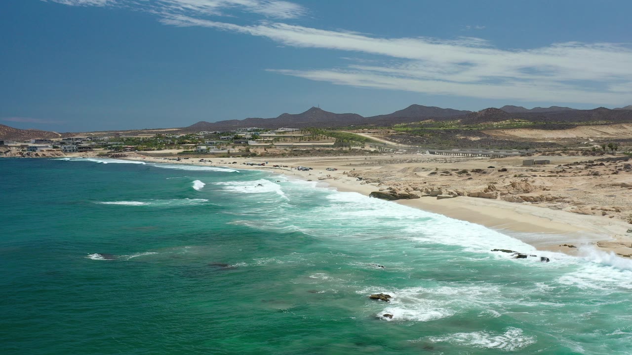 Wide aerial view of a Los Cabos beach with turquoise water in the foreground, white surf along the sand, dry terrain behind the shoreline, distant hills, and no visible people.
