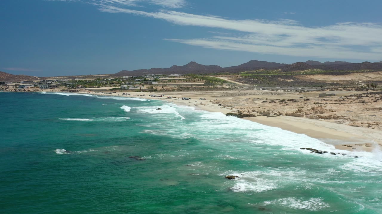 Drone-height beach view with ocean foreground, surf, sand, and mountains inland.