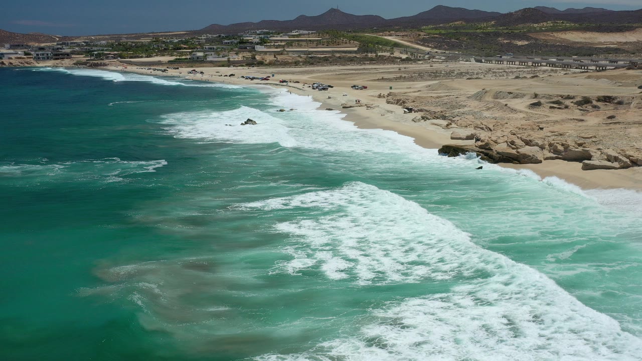 Aerial view over Los Cabos, Mexico with turquoise water and white surf breaking toward a sandy beach, rocky shoreline at right, low hills in the distance, and no visible people.