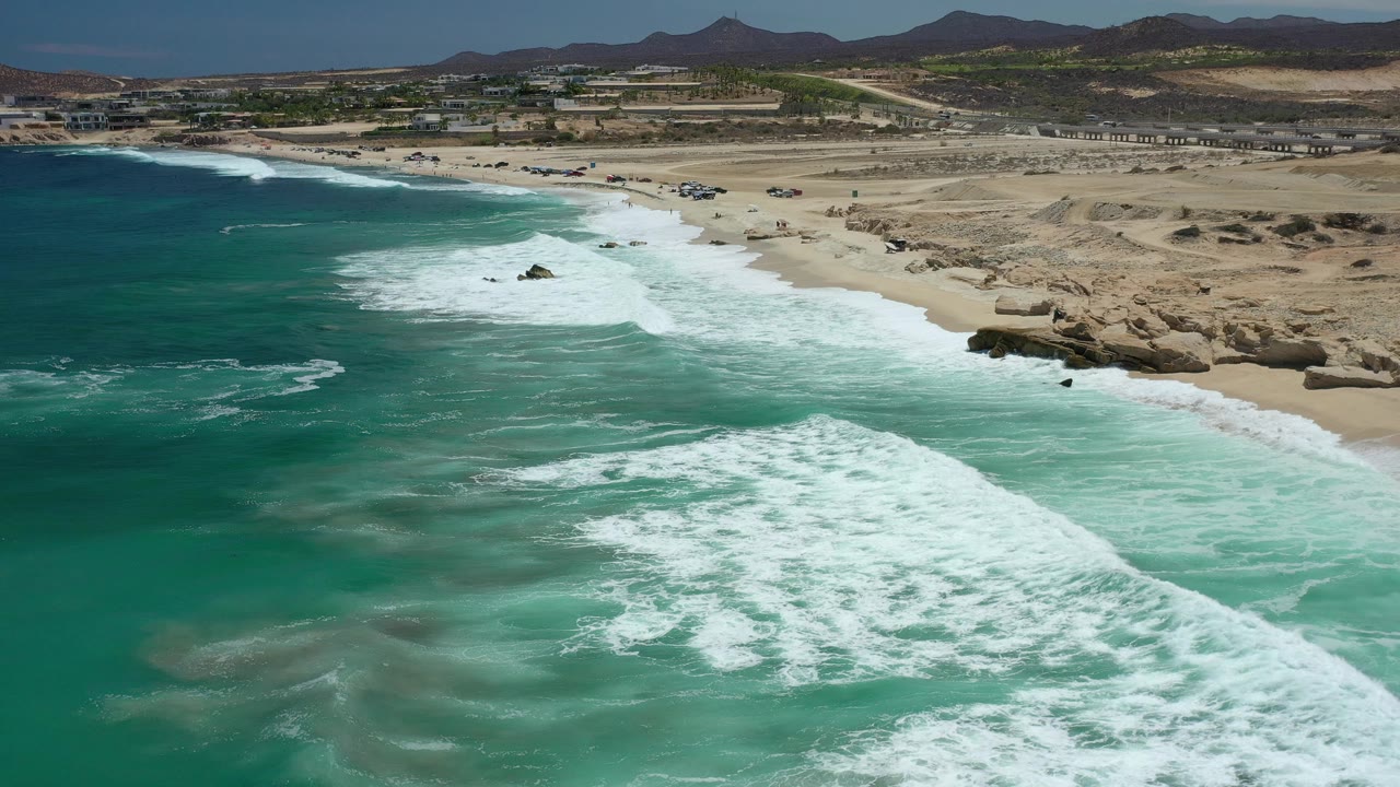 Aerial coastline with foamy surf and arid hills beyond the beach