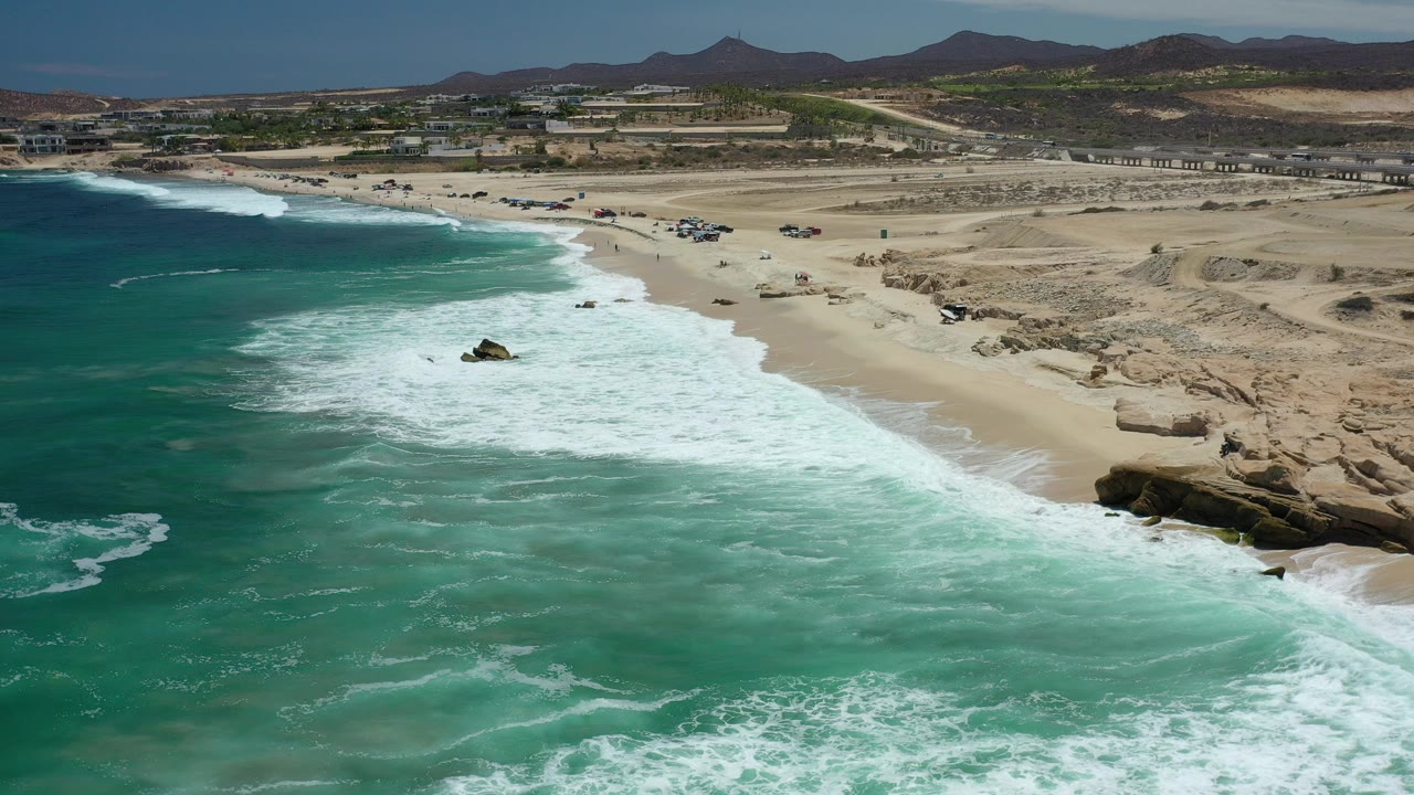 Aerial view of the Los Cabos coastline with turquoise water, white surf, a sandy barren beach, rocky outcrops, distant mountains, and no visible people at the opening moment.