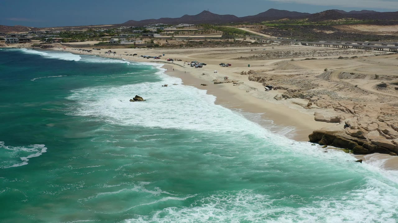 Aerial Los Cabos beach view showing foamy waves spreading across turquoise water, a pale sandy shore, exposed rocks at right, dry inland terrain, and no visible people.