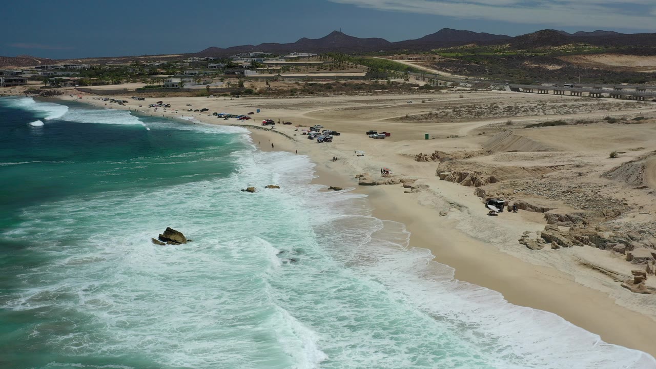 High aerial view of the Los Cabos coastline with turquoise surf on the left, pale sand beach, rocky shoreline, desert roads, distant mountains, and no visible people.